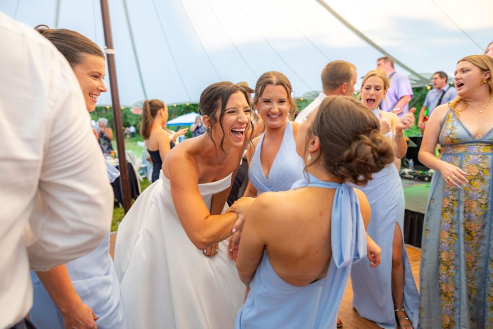 Bride laughing and hugging guests in light blue dresses at an outdoor wedding reception under a white tent.