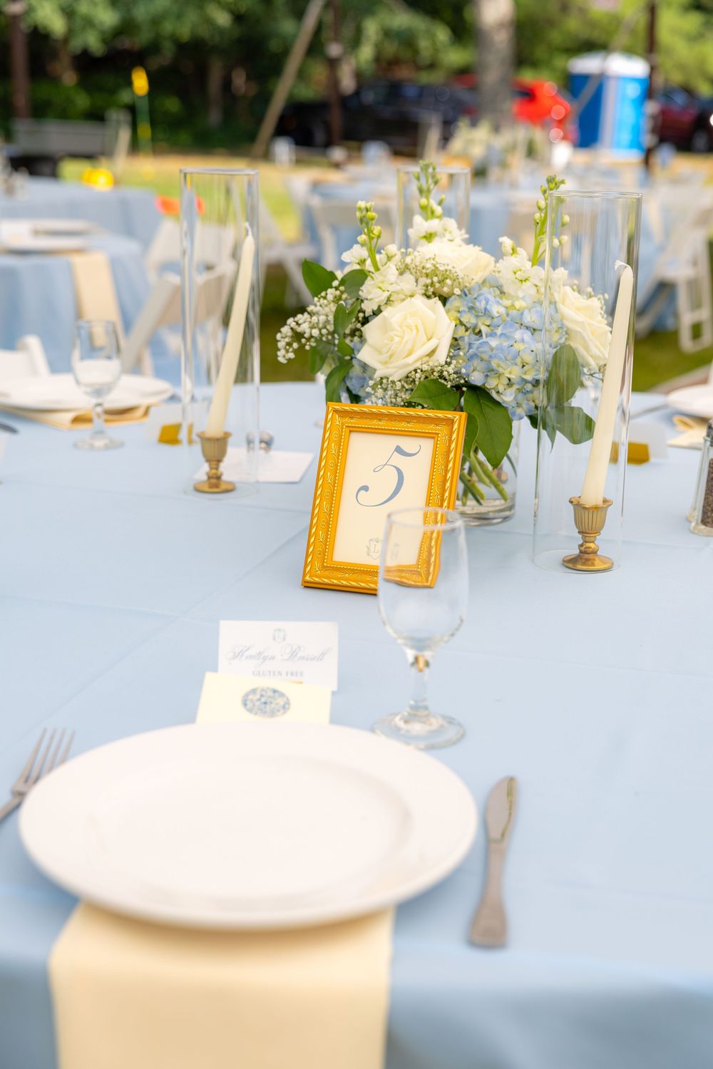 Elegant wedding reception table with blue linen, white plate, gold table number 5, and blue hydrangea floral centerpiece.