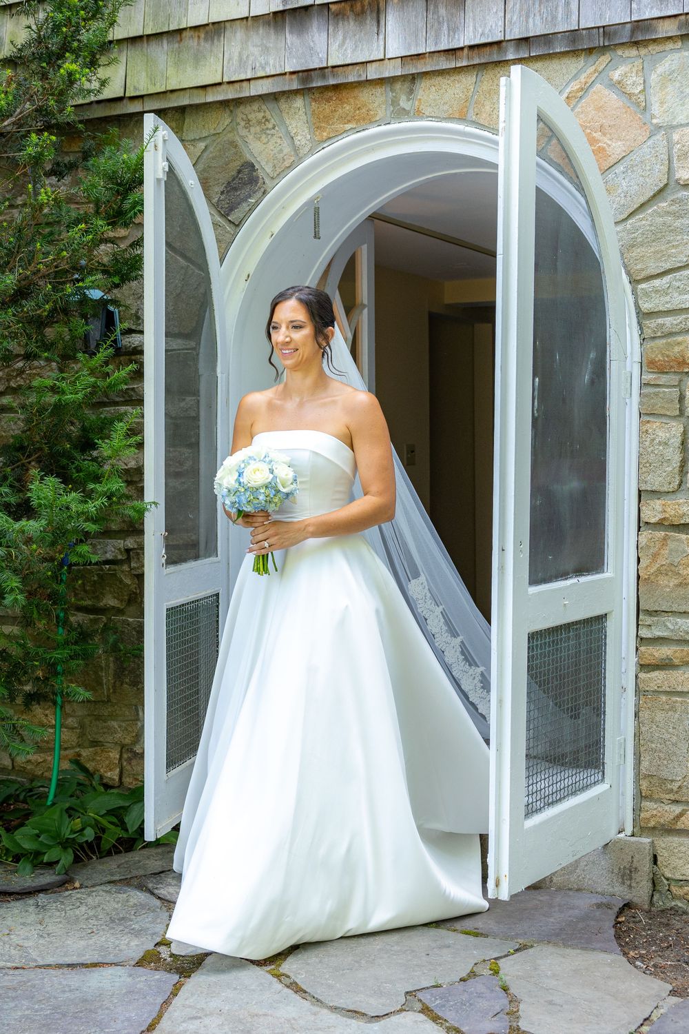 Bride in strapless white ball gown holding blue bouquet stands in arched doorway of stone building wearing veil.