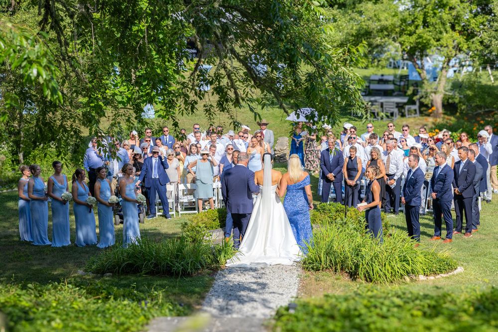 Outdoor wedding ceremony with bride, groom, and guests gathered under lush green trees.