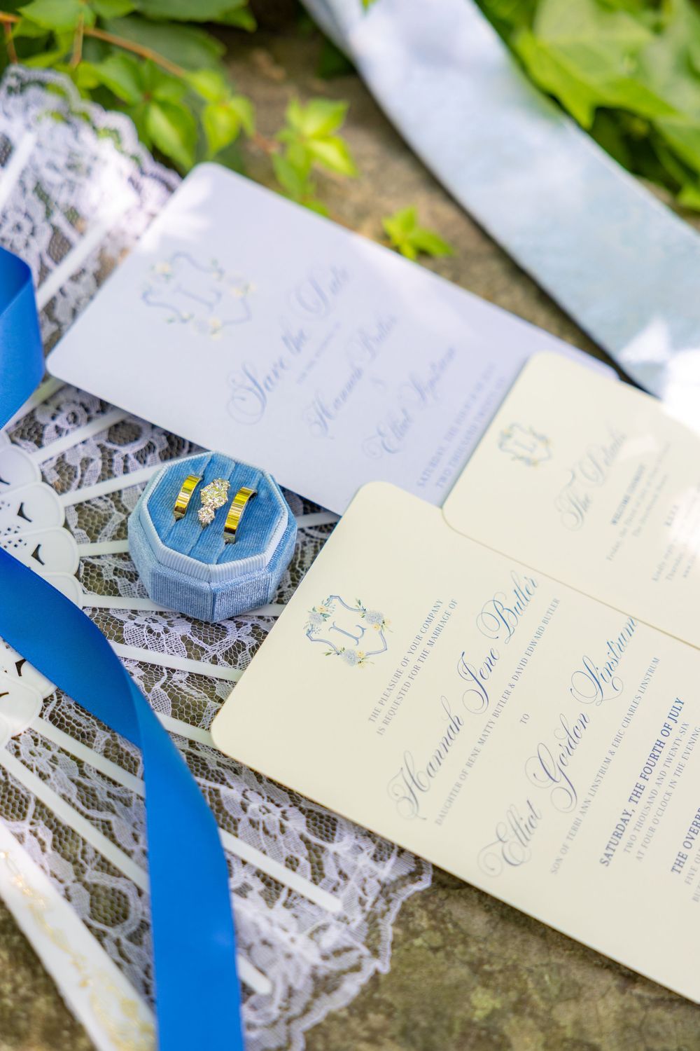 Elegant wedding invitation suite with blue ribbon, lace, ring box with gold bands, and cream stationery on stone surface.