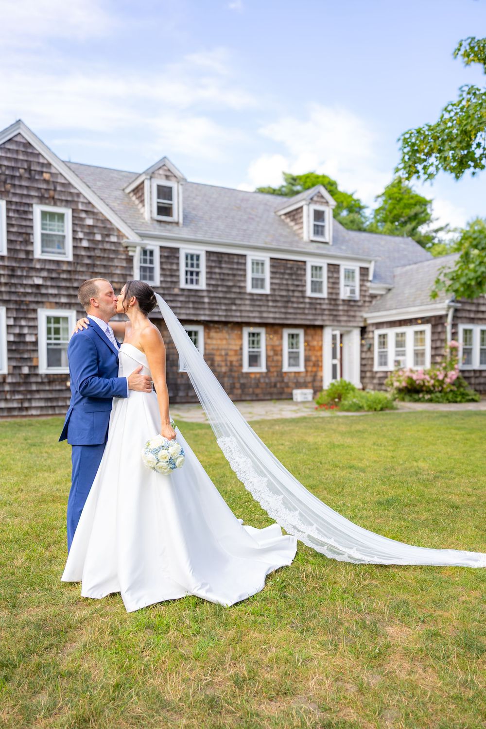 Bride and groom share a kiss on green lawn in front of classic cedar shingle estate, her lace veil flowing in the breeze.
