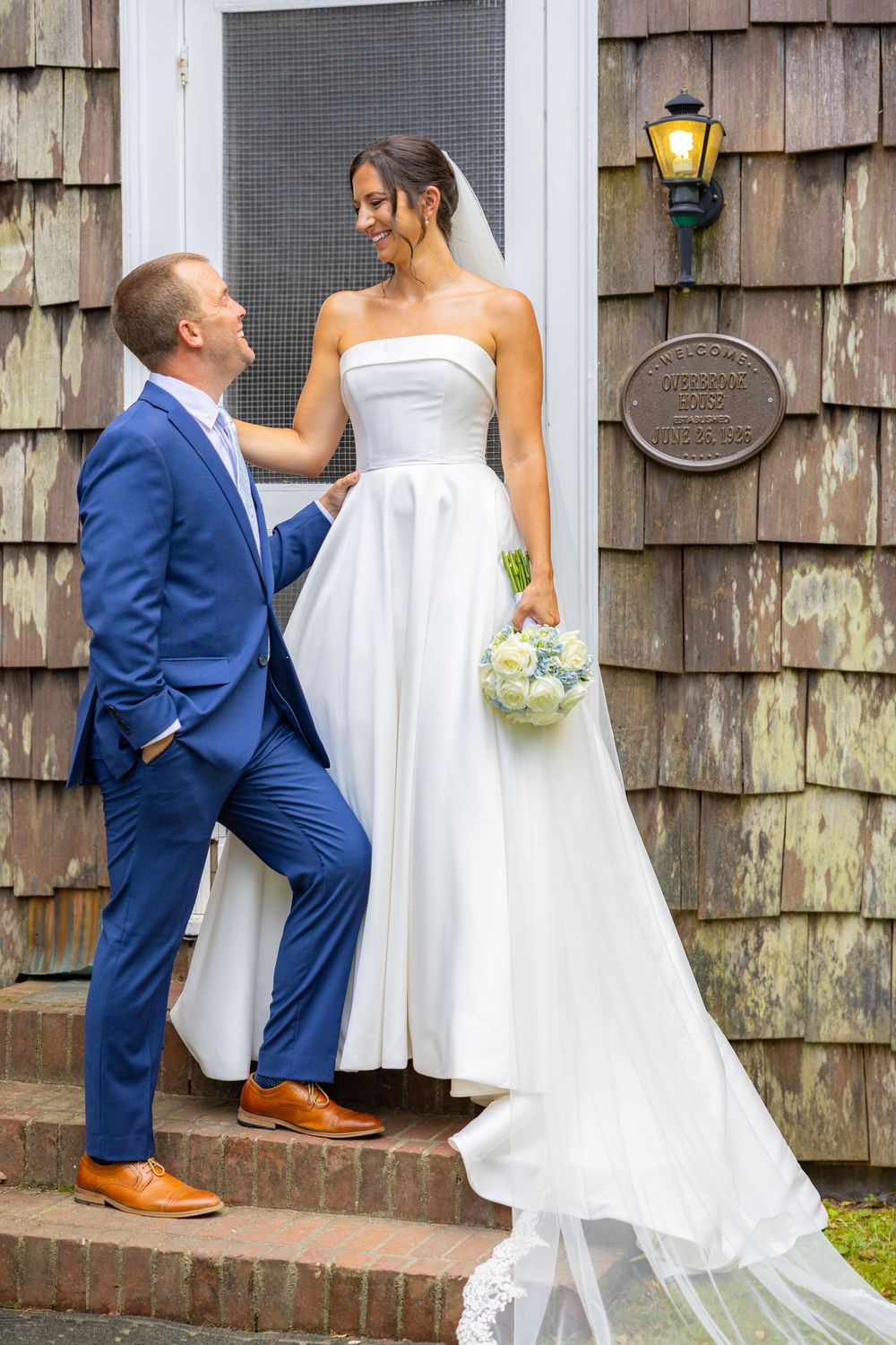 Bride in white strapless gown holding bouquet stands on steps with groom in blue suit outside rustic shingle building.