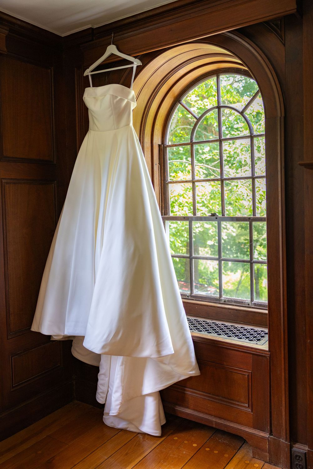 White strapless wedding gown hanging on hanger in front of arched window in elegant wood-paneled room.
