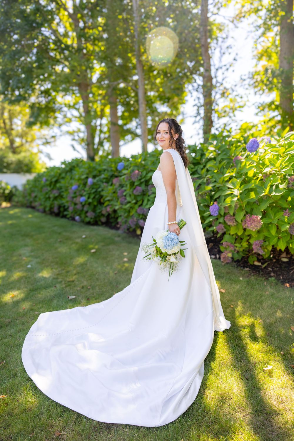 Bride in white gown with long train holds blue hydrangea bouquet in lush garden setting.