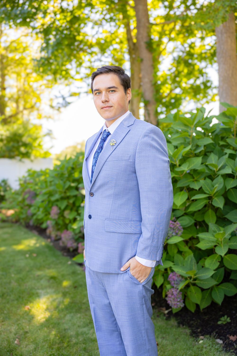 A man in a light blue suit and blue tie stands outdoors among green hydrangea bushes at a wedding.