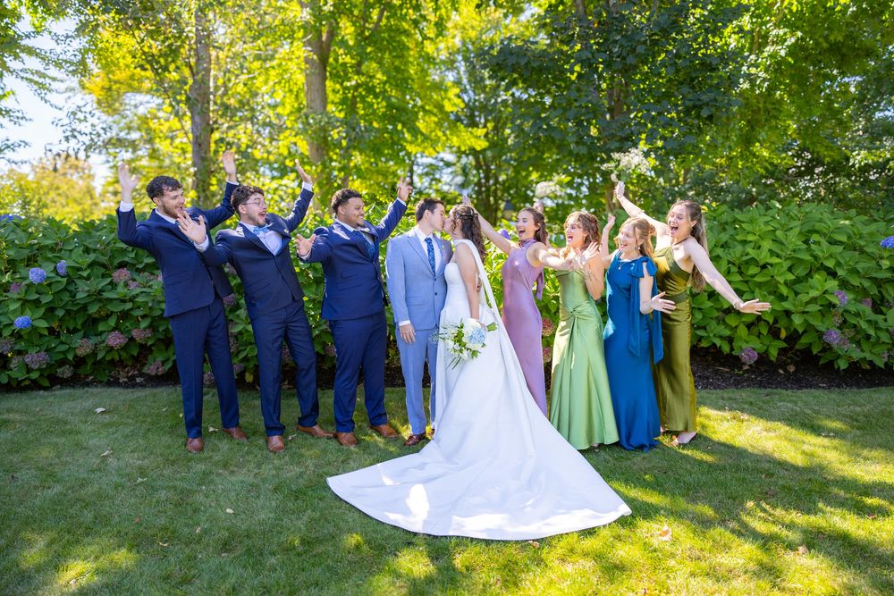 Wedding party celebrating outdoors on green lawn, bride in white gown with groomsmen in navy suits and bridesmaids in colorful dresses.