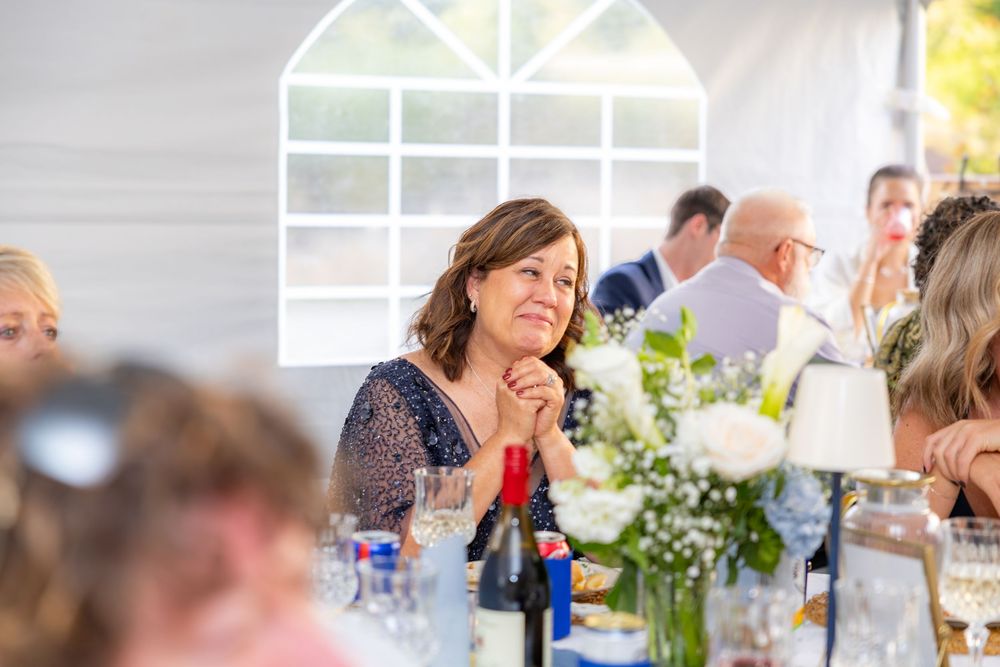 A woman smiles warmly at a wedding reception, seated at a decorated table under a white marquee tent.