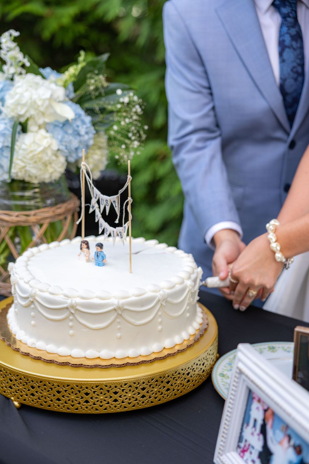 A couple cuts a white wedding cake with figurine toppers on a gold stand at an outdoor ceremony.