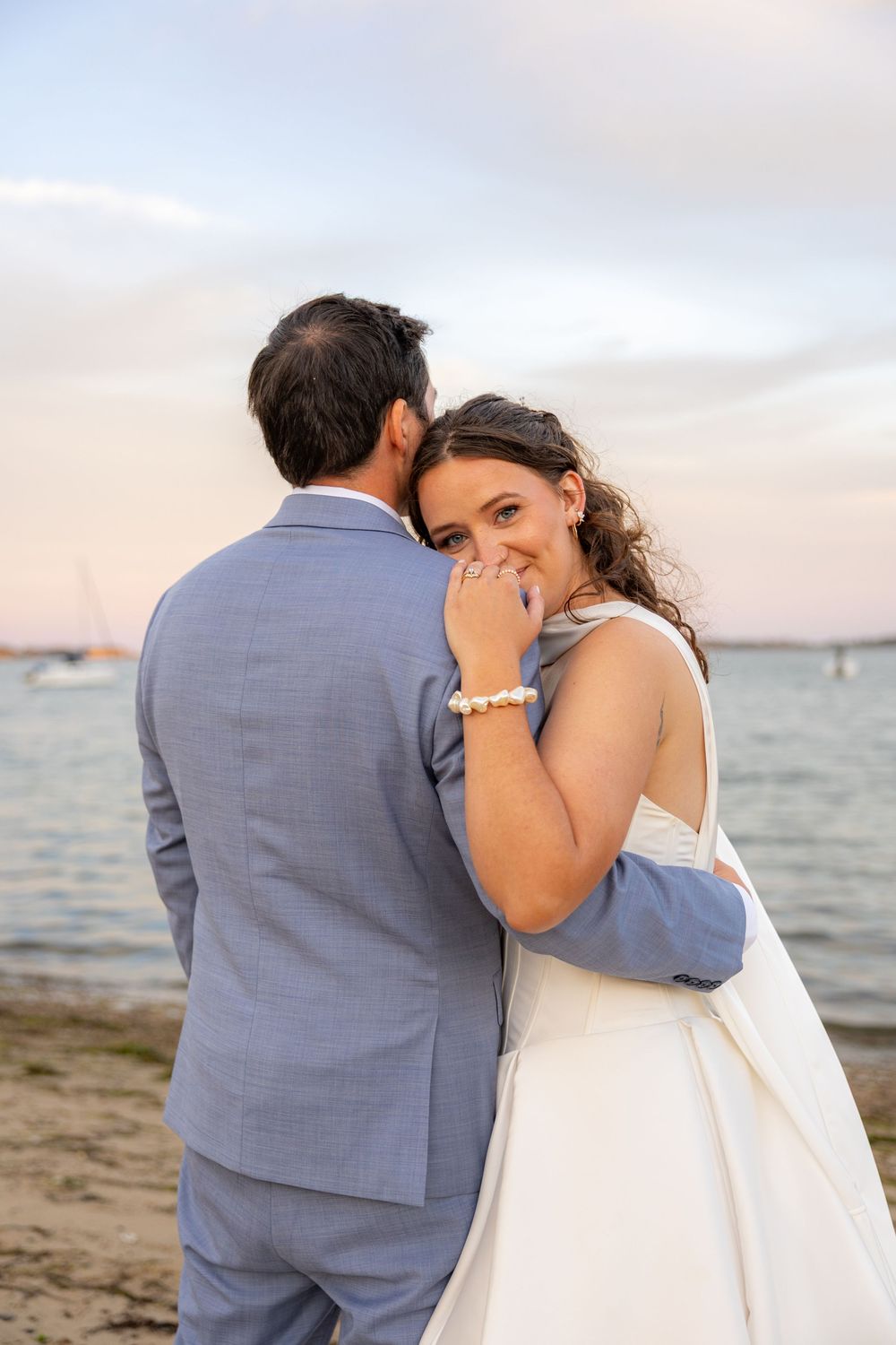 Bride in white dress smiles joyfully as groom in blue suit whispers to her on a sandy beach at sunset.