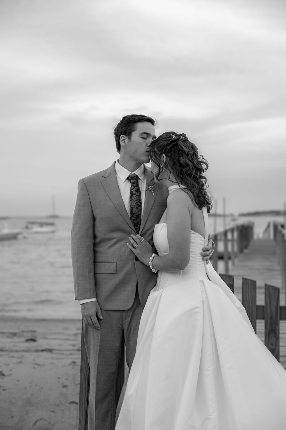 Black and white photo of a bride and groom sharing a kiss on a beach boardwalk on their wedding day.