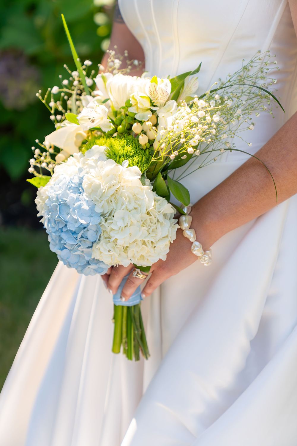 Bride in white gown holding a wedding bouquet of blue hydrangeas, white roses, and baby's breath outdoors.
