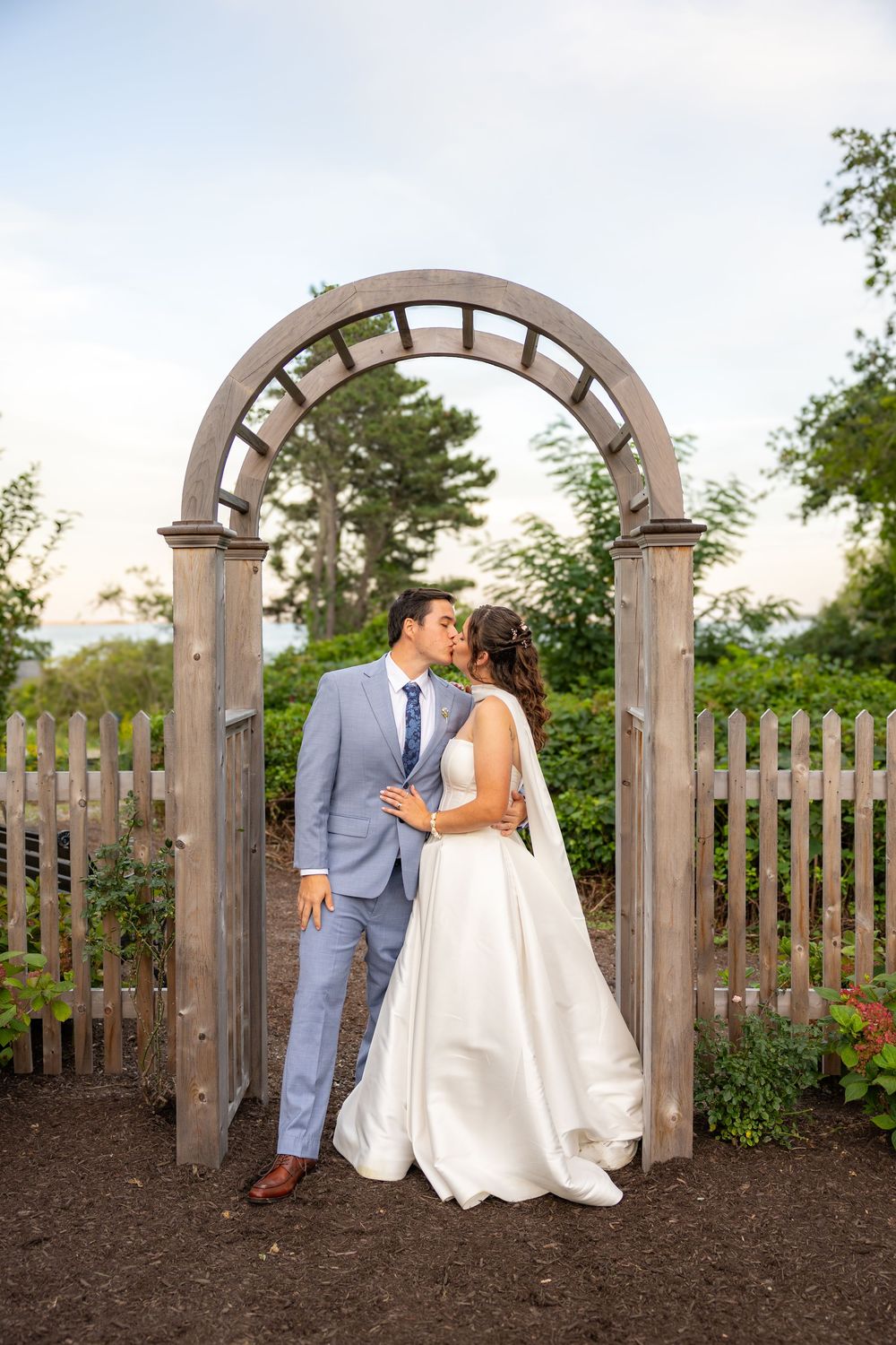 Bride and groom share a kiss under a rustic wooden arch in a garden wedding setting.