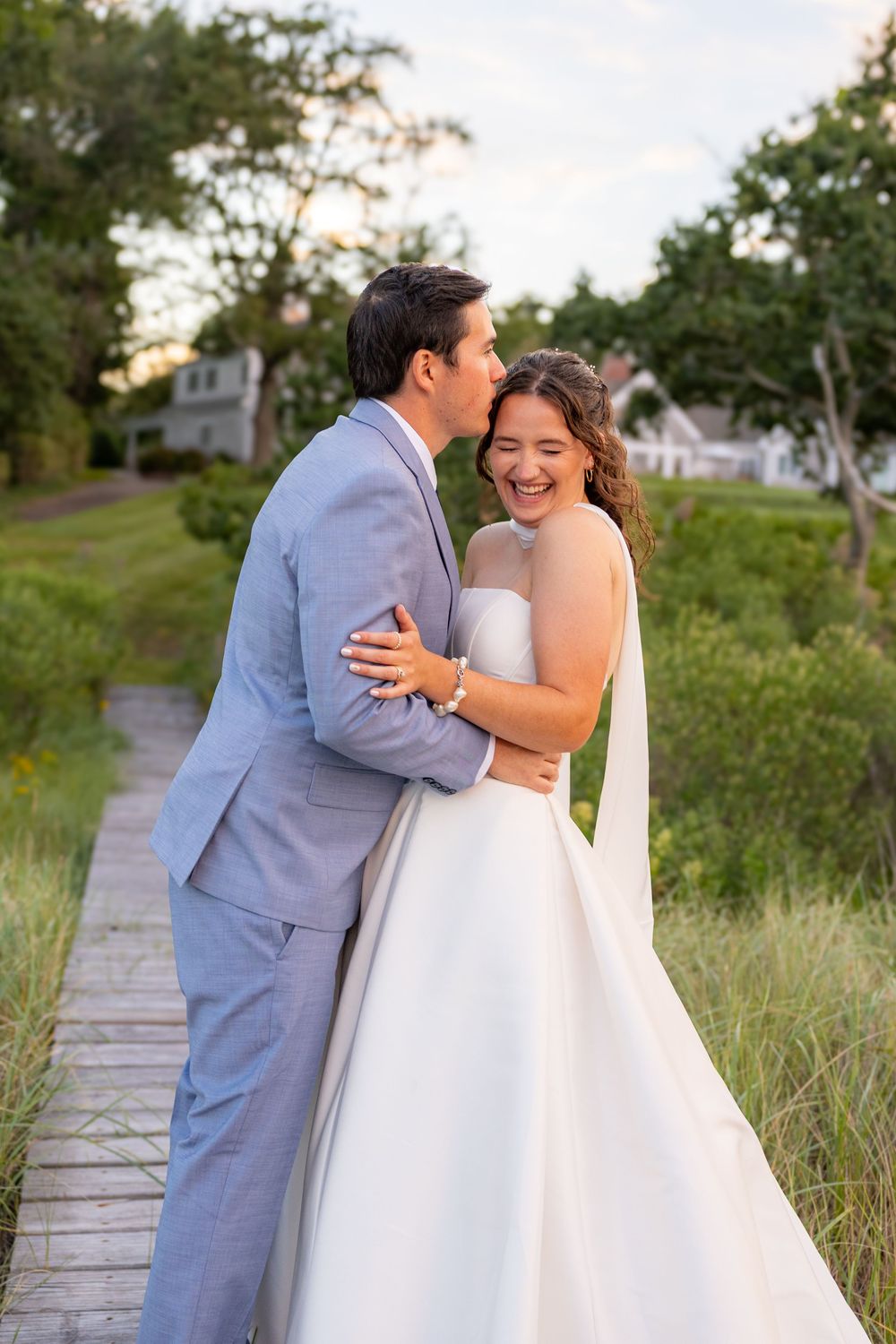 A joyful bride and groom embrace on a wooden path, surrounded by lush greenery, as the groom kisses her cheek.