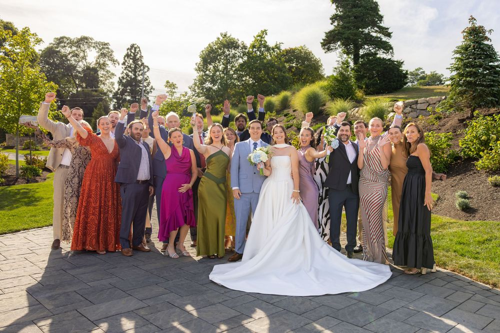 Wedding couple poses with guests outdoors at garden venue on sunny day.