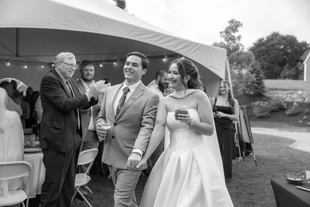 Black and white photo of a smiling bride and groom walking hand in hand at an outdoor wedding reception.