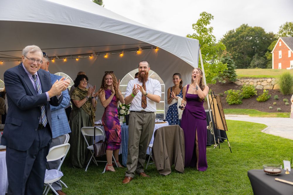 Guests clapping and celebrating outdoors at a wedding reception under a white tent with string lights.