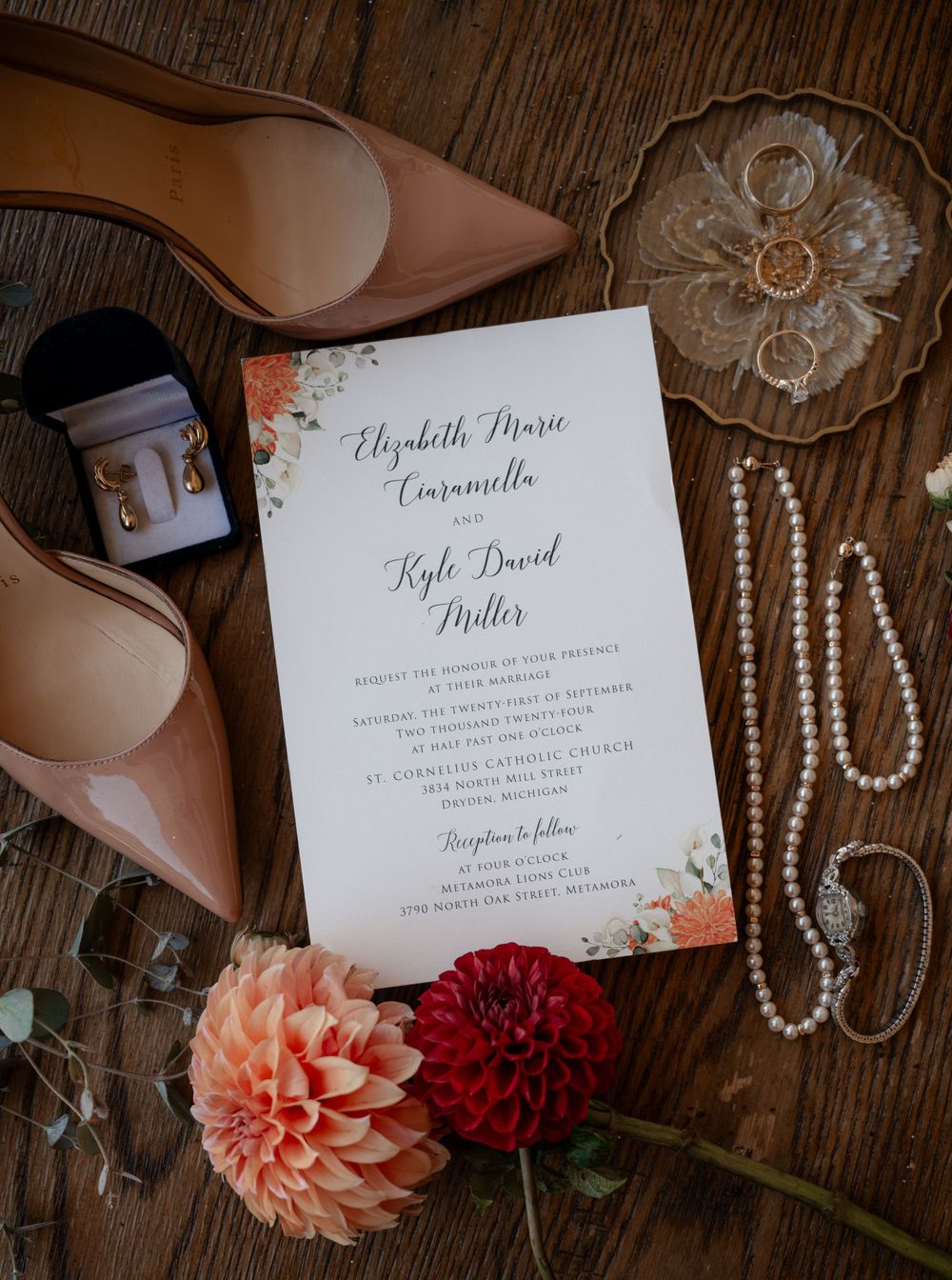 Wedding invitation for Elizabeth Marie Carravella and Kyle David Miller surrounded by heels, pearls, earrings, and dahlia flowers.