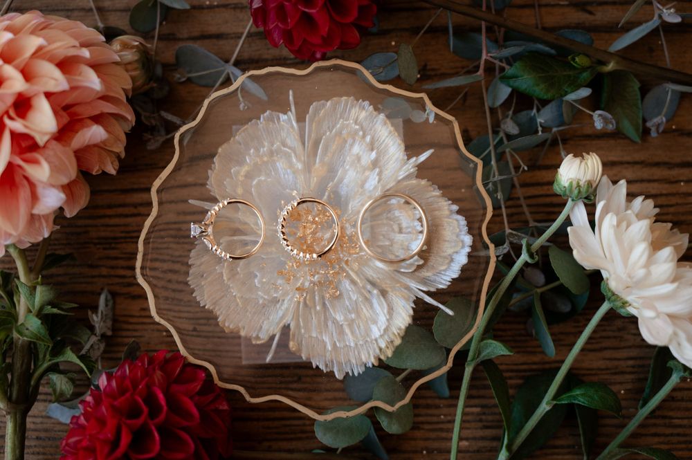 Gold wedding rings displayed on a white feather dish surrounded by pink and red dahlia flowers on a wooden surface.