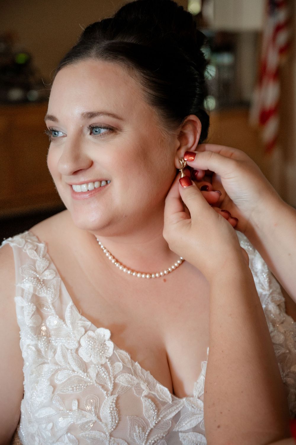 Smiling bride in lace wedding gown wearing pearl necklace while having gold earrings put on.