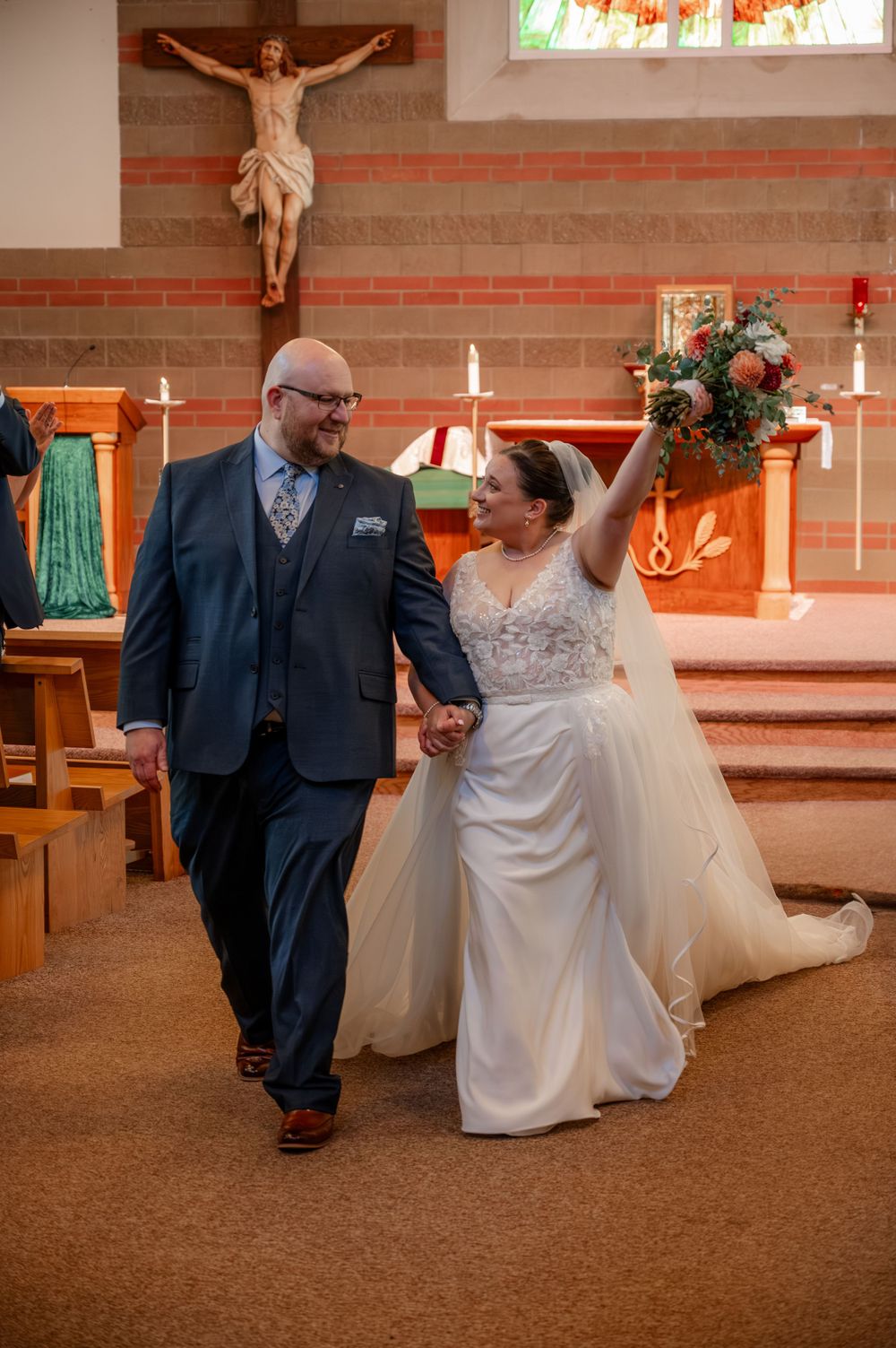 Newlyweds walking down church aisle after ceremony, bride joyfully raising her bouquet beneath a crucifix.