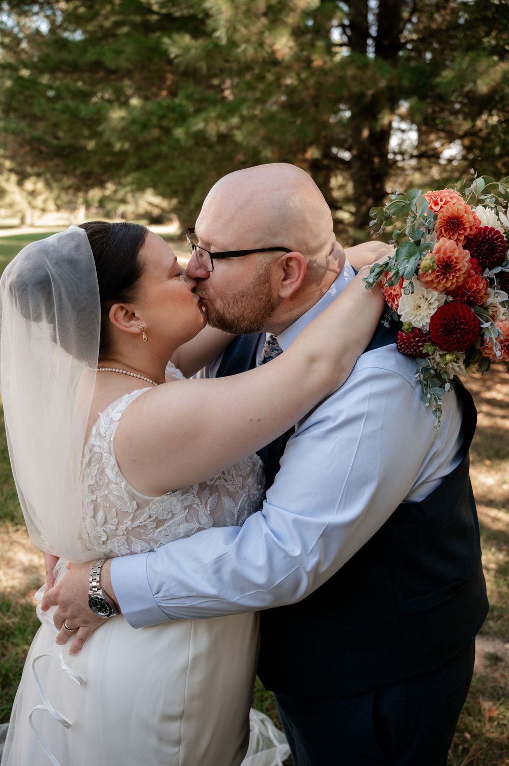 Bride and groom sharing a kiss outdoors, bride holding a red and coral dahlia wedding bouquet.