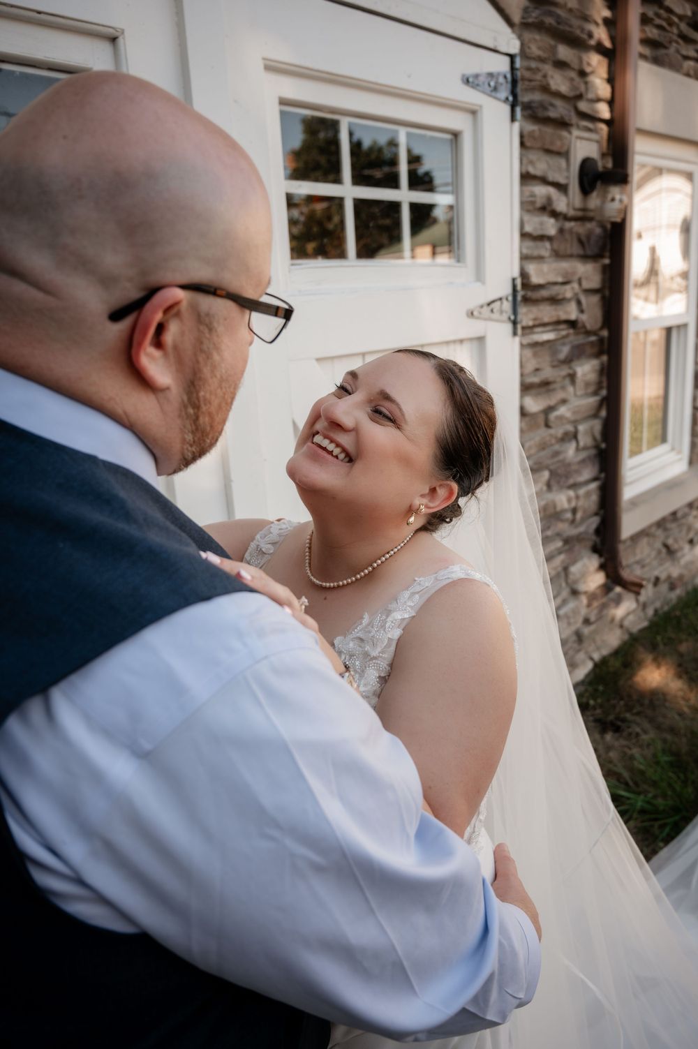 Bride in lace gown and veil smiling up at groom outside a stone building in golden hour light.