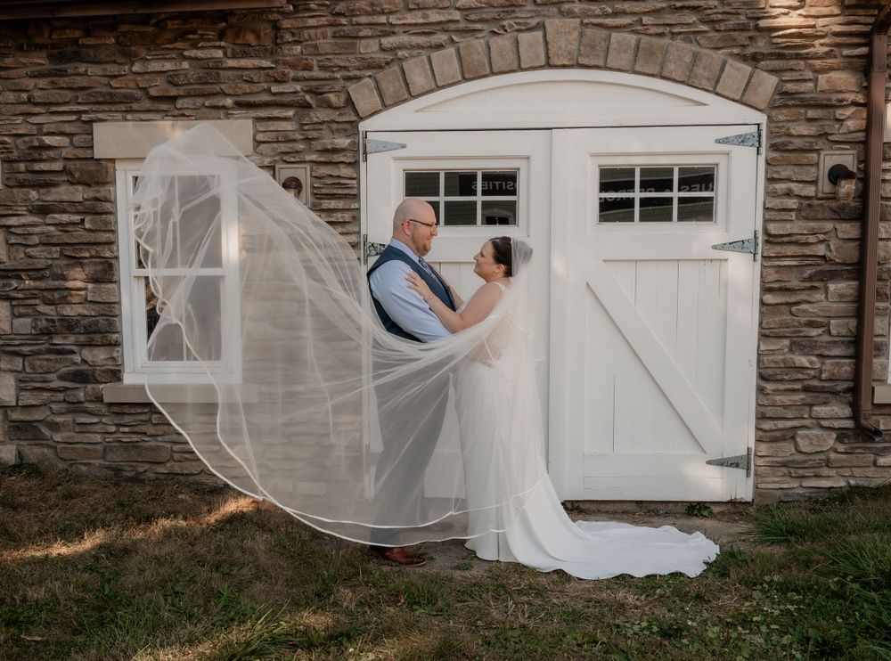 Bride and groom embracing in front of white barn doors as the wedding veil billows in the wind.
