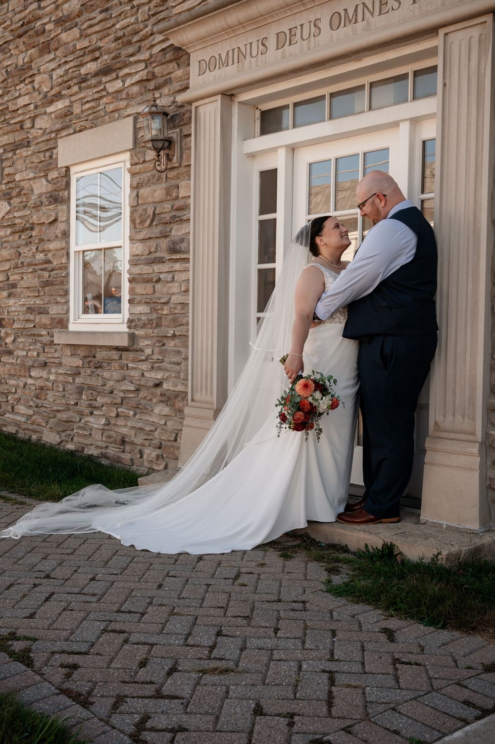 Bride and groom sharing a tender moment outside a stone building with Latin inscription above the door.
