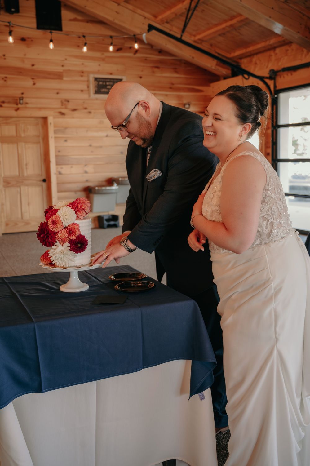 Smiling bride and groom cutting their elegant dahlia-adorned wedding cake in a rustic barn venue.