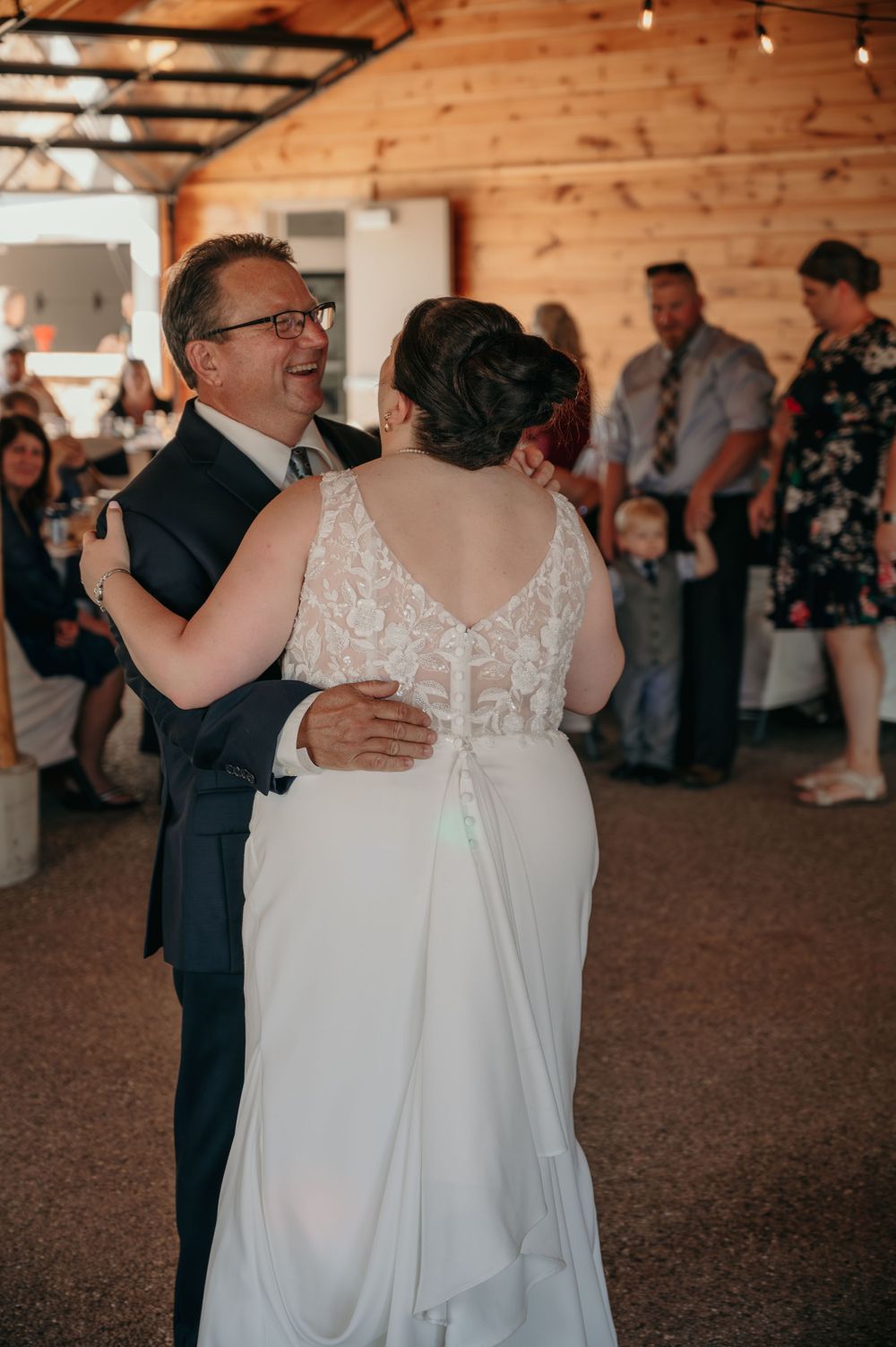 Groom smiles joyfully while dancing with bride in lace wedding gown at barn reception.
