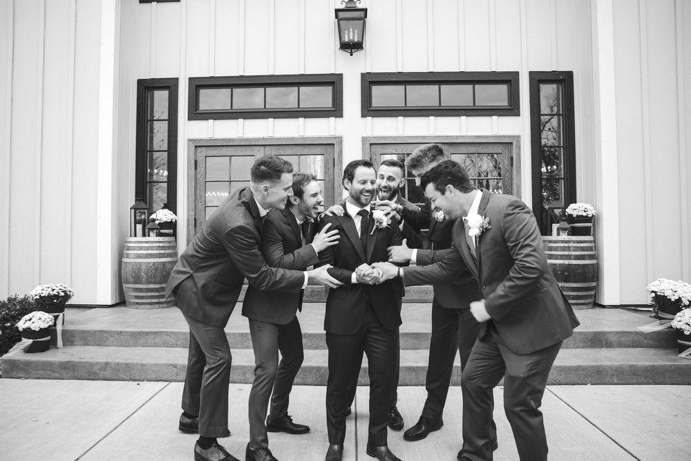 Groom and groomsmen laughing together outside a white barn venue in matching dark suits.