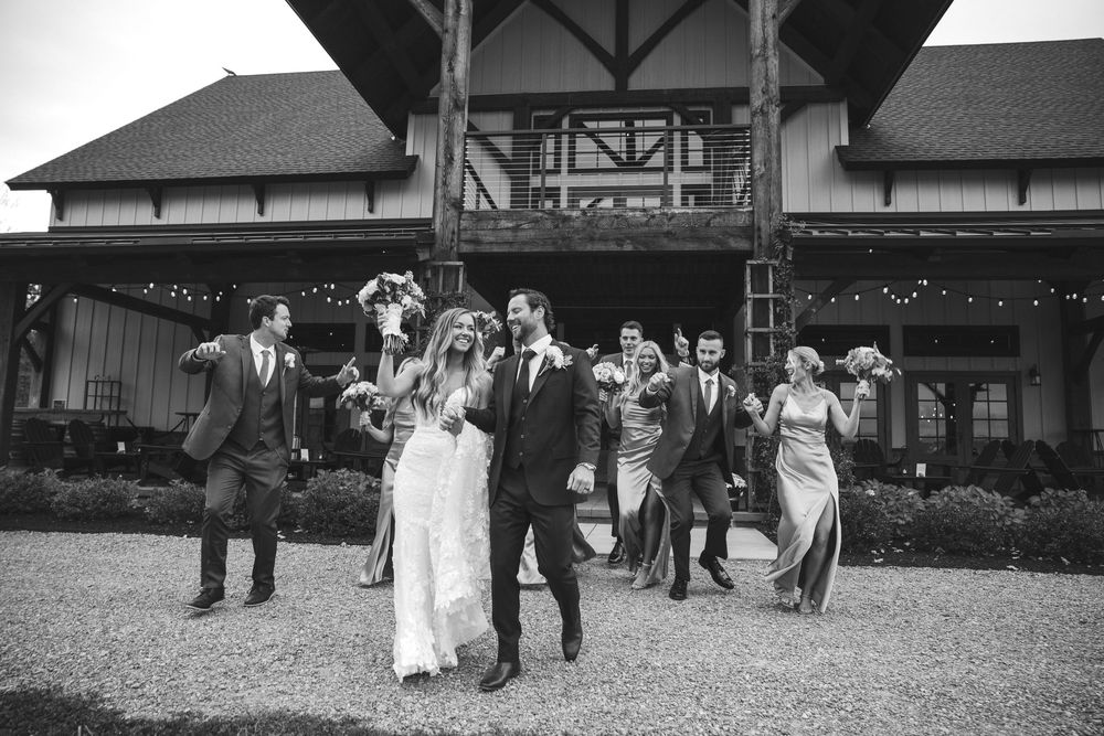 Bride and groom lead wedding party in joyful walk outside rustic venue in black and white photo.