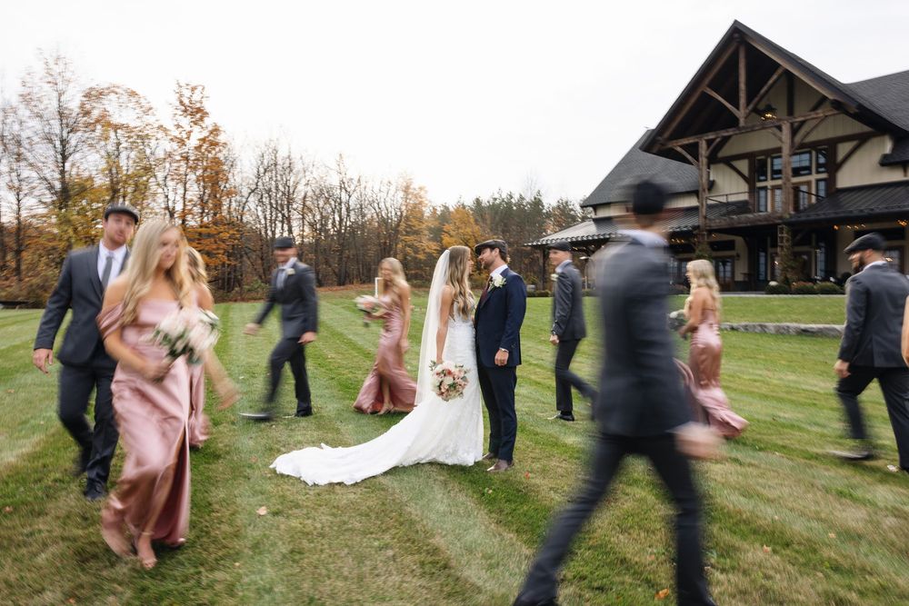 Bride and groom stand together as wedding party walks past rustic lodge venue in autumn setting.
