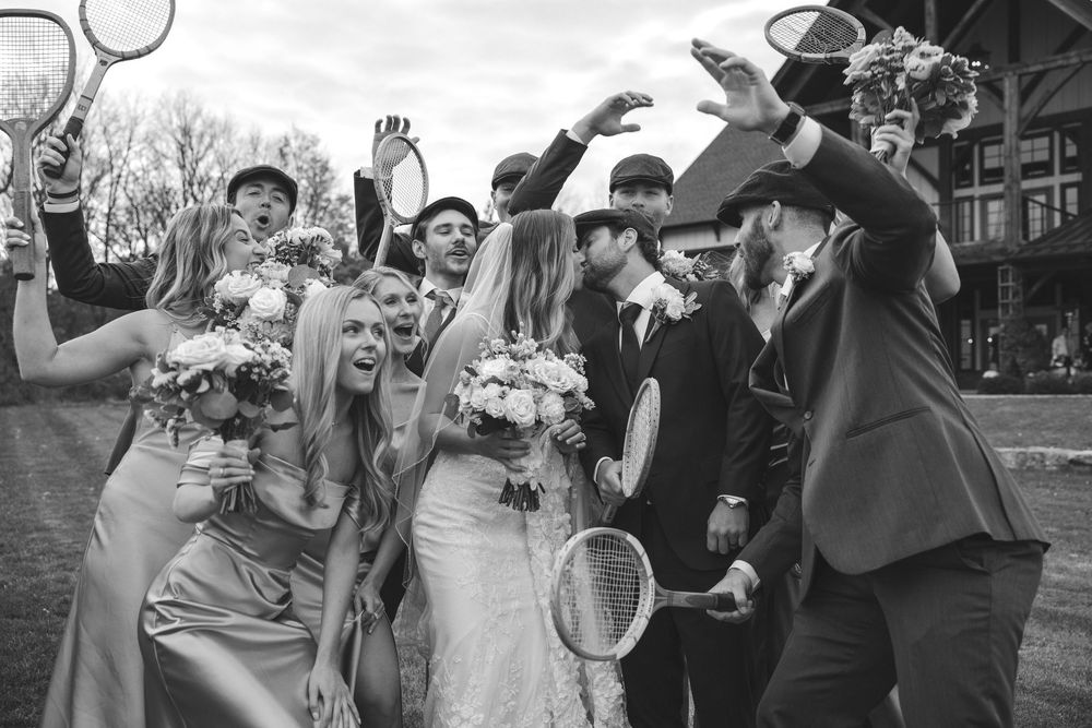 Laughing wedding party poses with tennis rackets outside rustic venue in fun black and white photo.