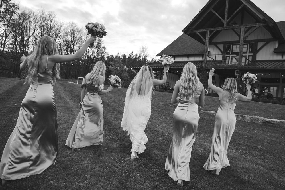 Bride and bridesmaids in elegant gowns walk across lawn raising bouquets outside wedding venue.