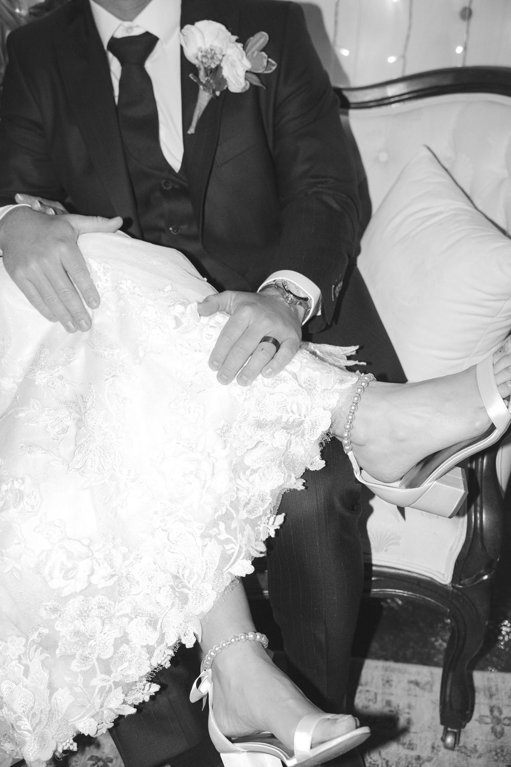 Close-up of bride in lace gown and groom in tuxedo seated together showing wedding rings and heels.