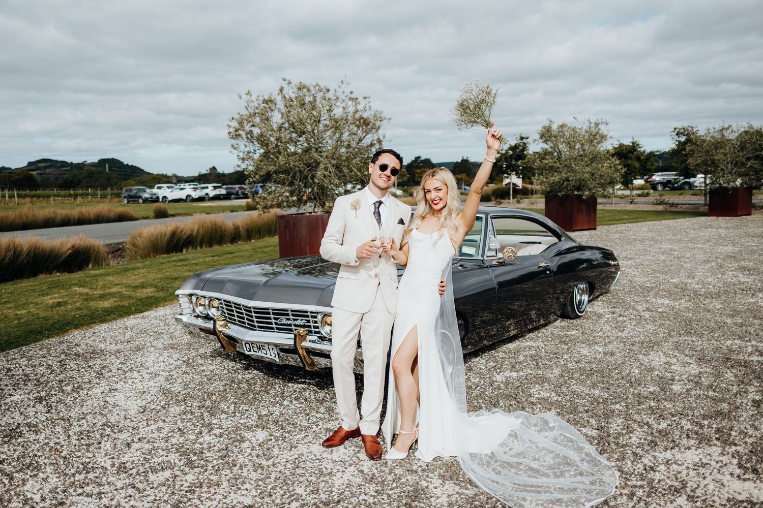 A celebratory wedding photo featuring a classic Chevrolet car and vineyard backdrop.