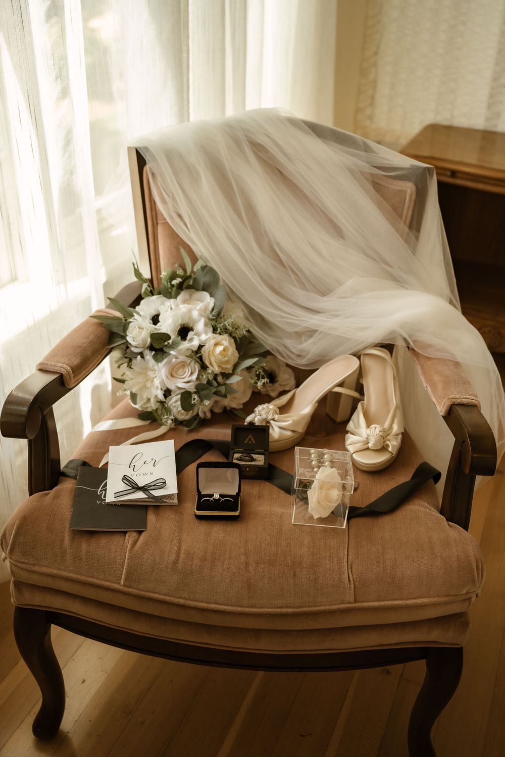 Wedding dress and floral bouquet elegantly draped over a brown chaise lounge in natural window light.