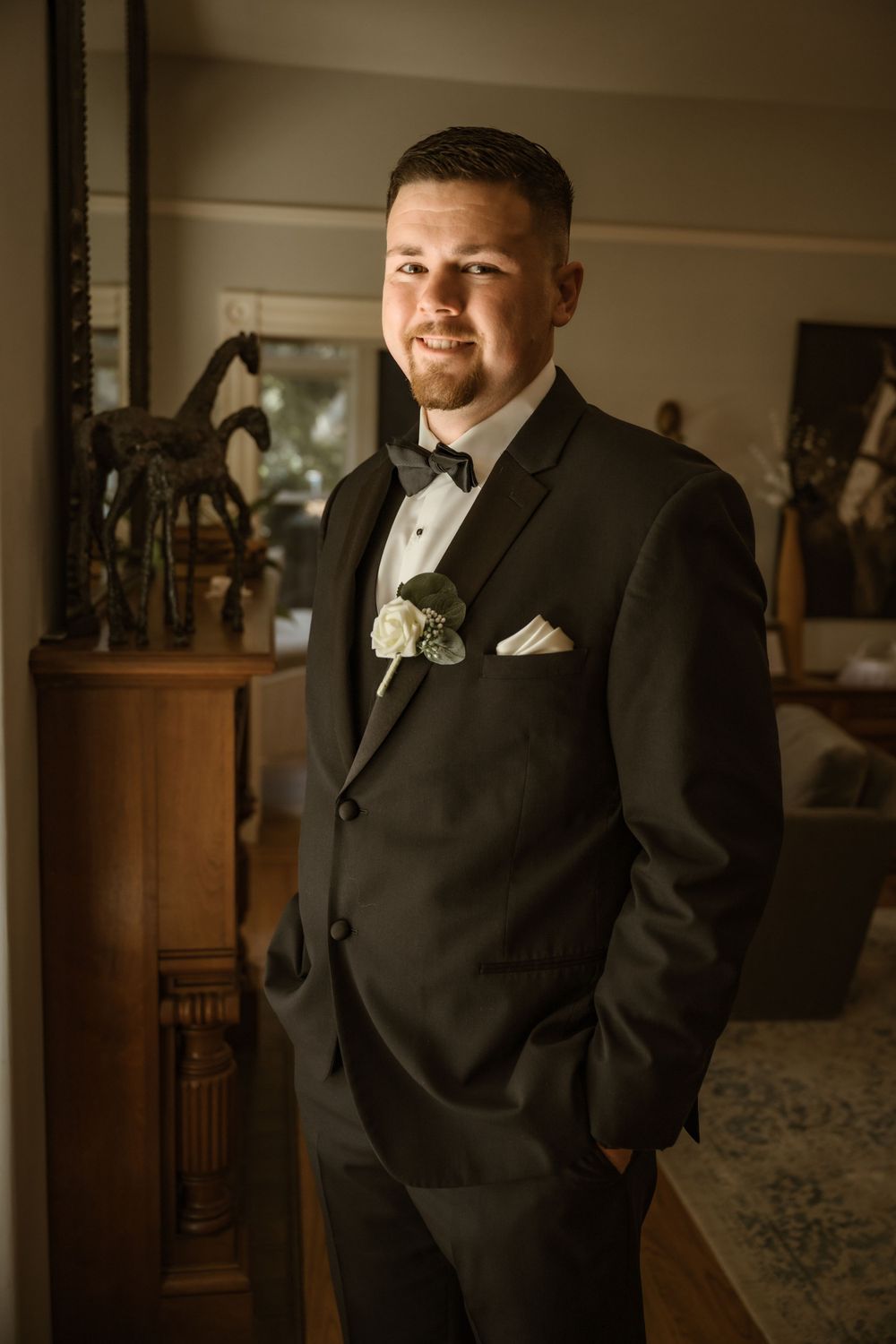 Formal portrait of groomsman in black tuxedo with white boutonniere against dark background.