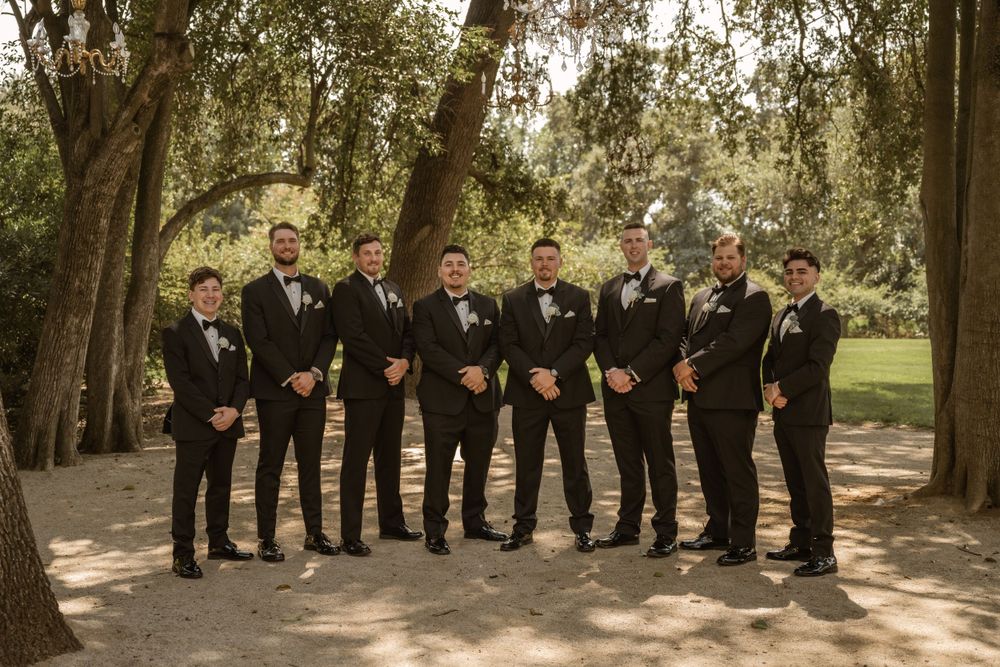 Large wedding party in black tuxedos posed in line under trees outdoors.