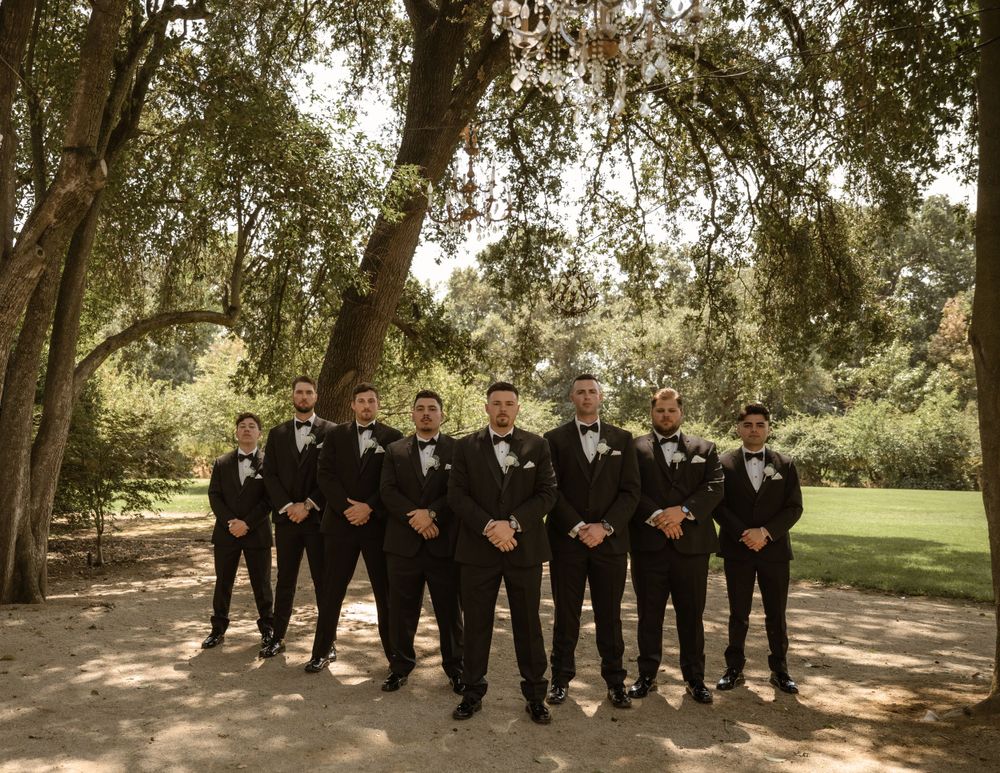 Groomsmen in black suits standing in formation under tree canopy at outdoor venue.