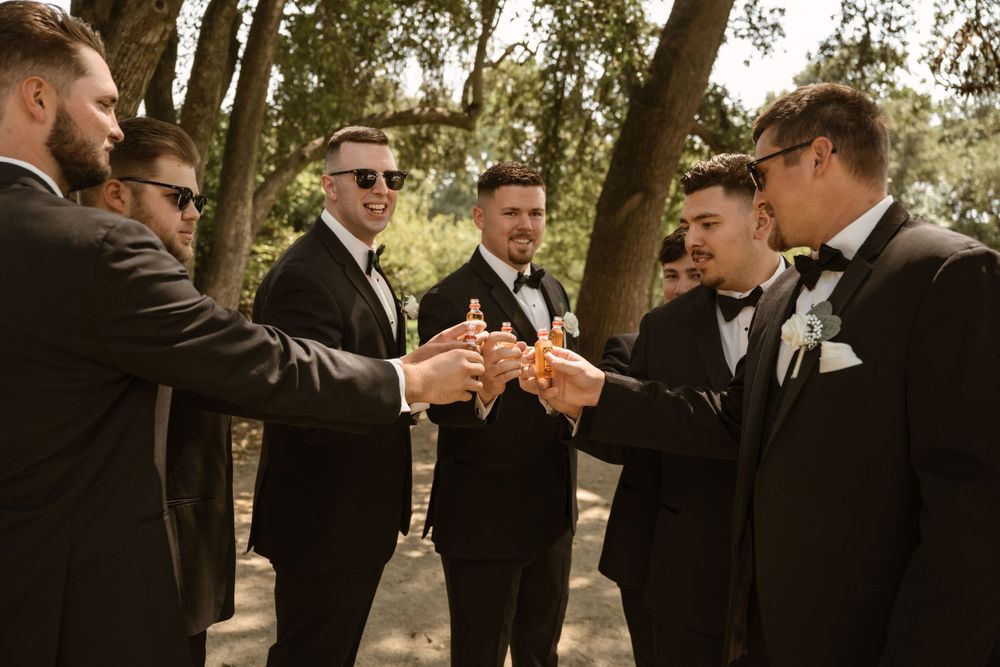 Wedding party in formal attire toasting with drinks outdoors under trees.
