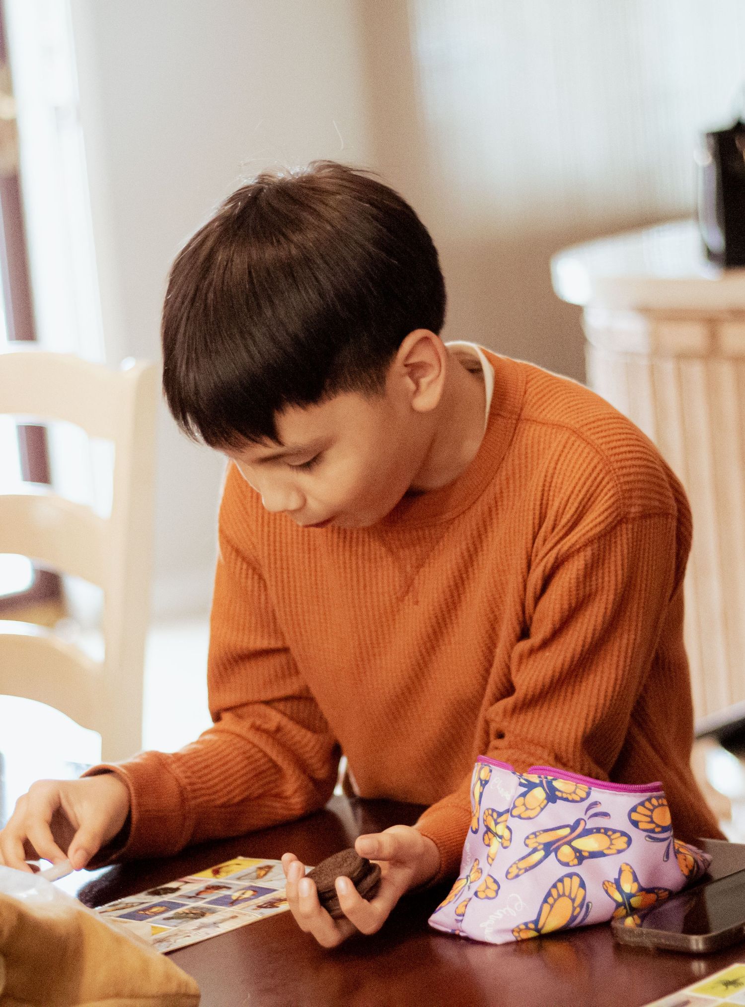 Young person in orange sweater focuses intently on reading or writing at a table.