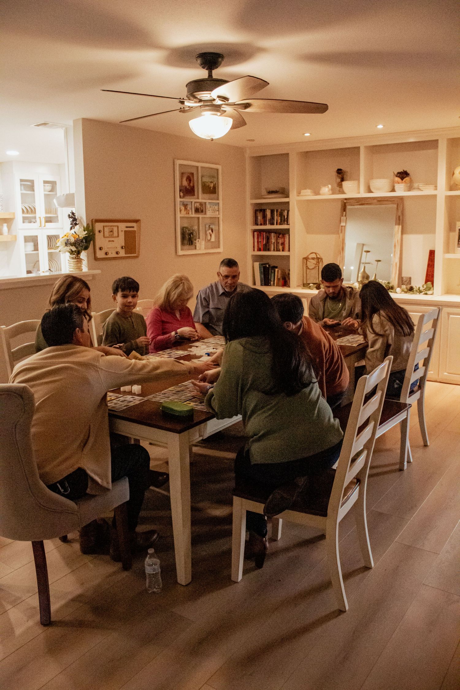 Large group gathered around a dining table playing games in a warmly lit room with built-in bookshelves.