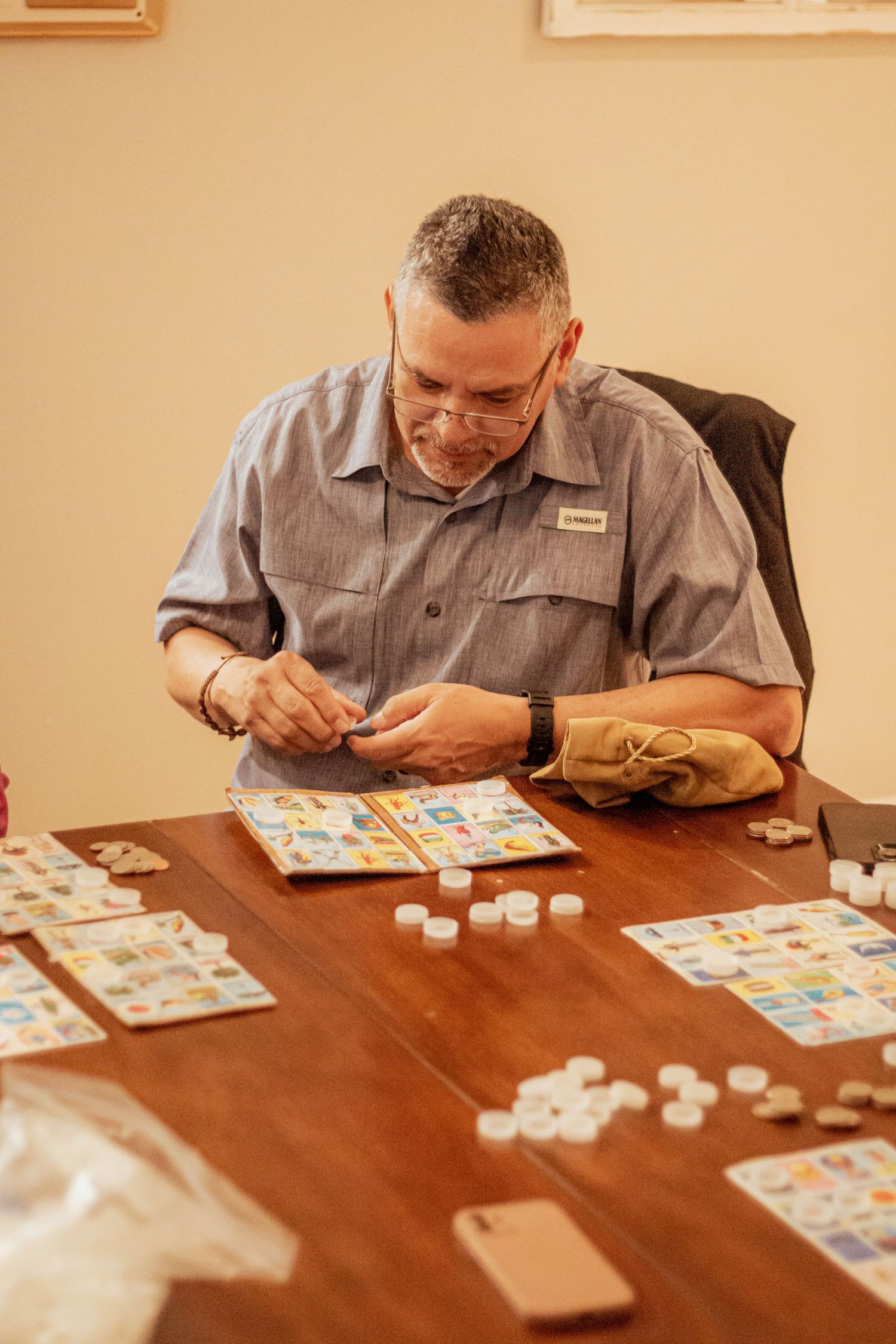 Someone wearing glasses and gray work shirt concentrating while playing a board game with tiles at wooden table.