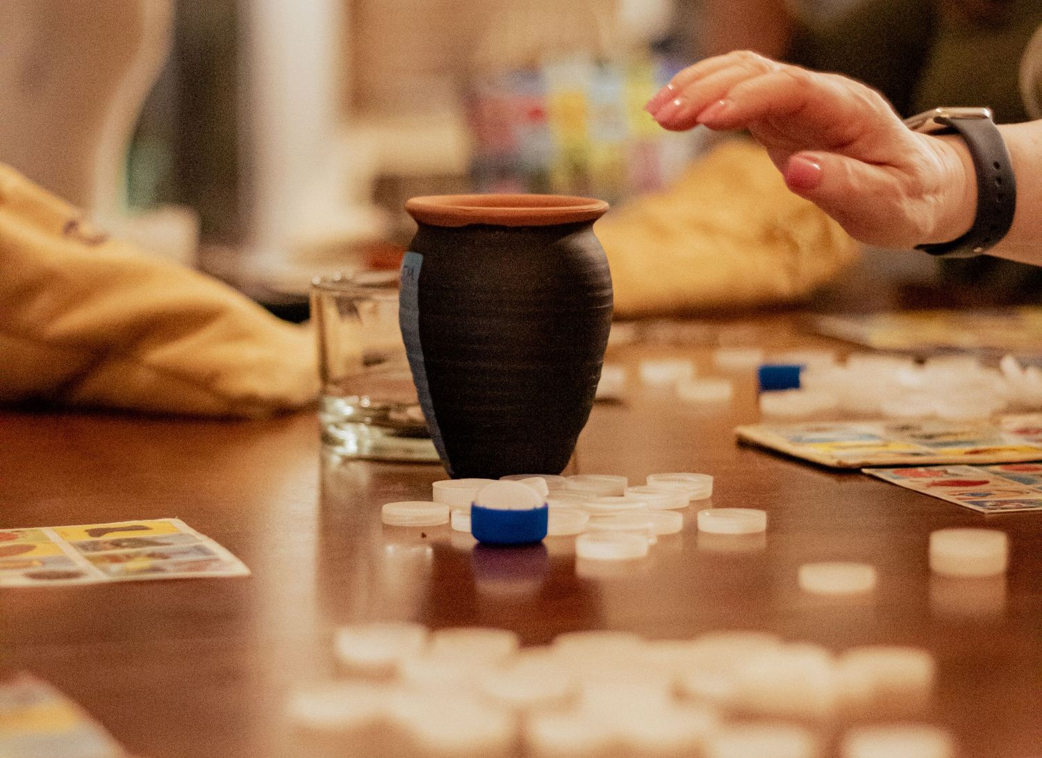 Playing cards and a dark ceramic jar sit on a wooden table during a tabletop gaming session.