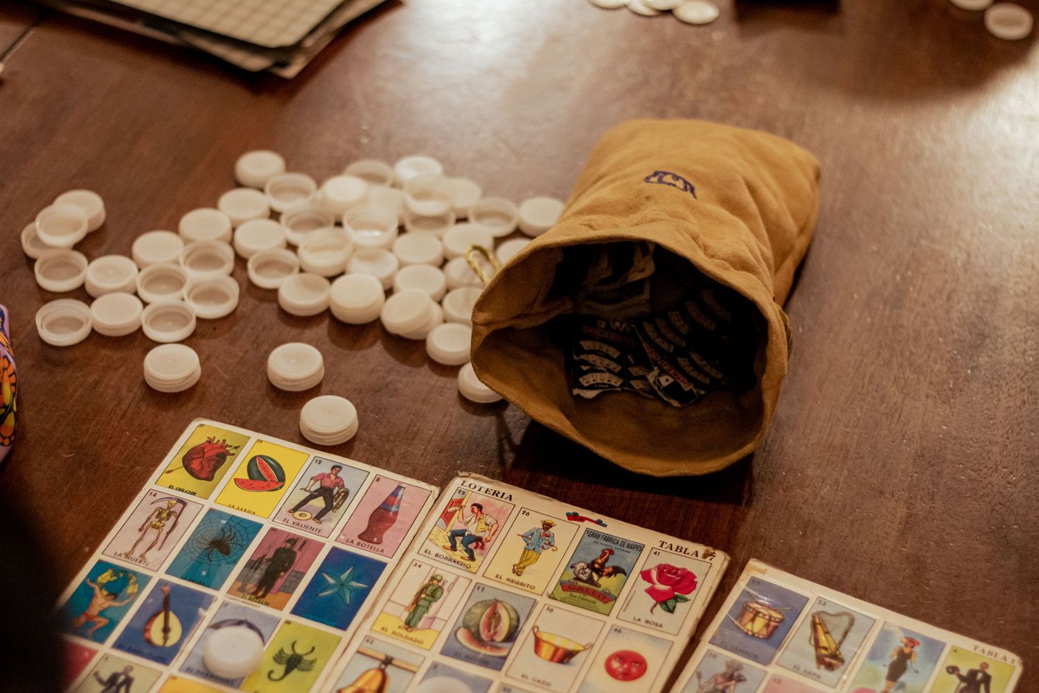 Mexican Loteria cards laid out on a wooden table with white circular game tokens and a brown burlap bag.