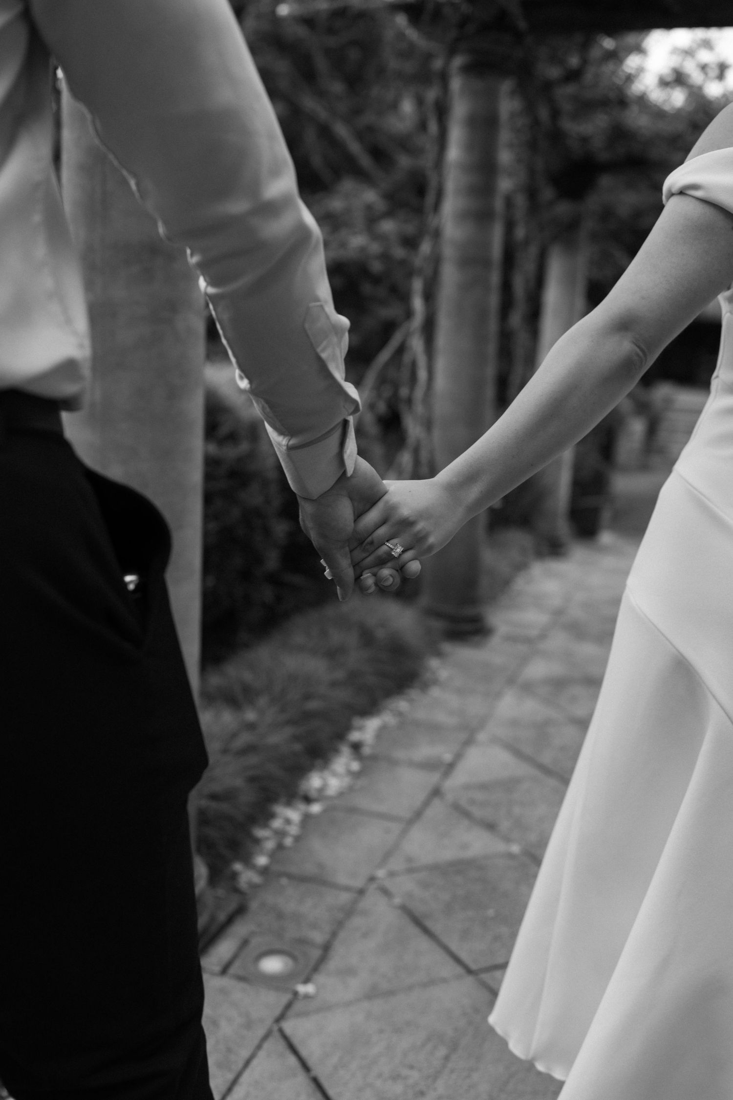 A black and white photo of two people holding hands on a garden path.
