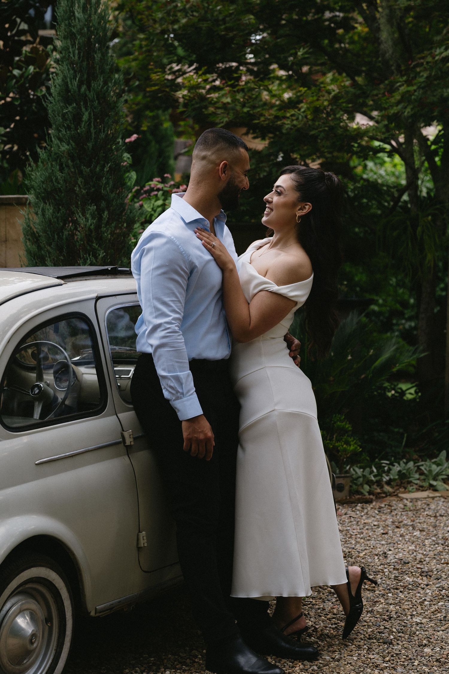 A couple in formal attire poses romantically next to a vintage car in a garden setting.